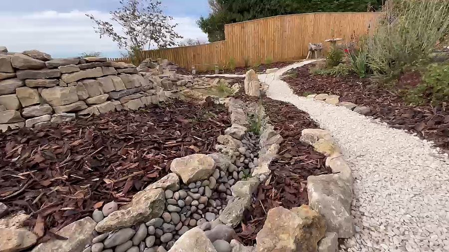 A pebble-lined rill running between drystone edges, beside a gravel path and a long drystone wall