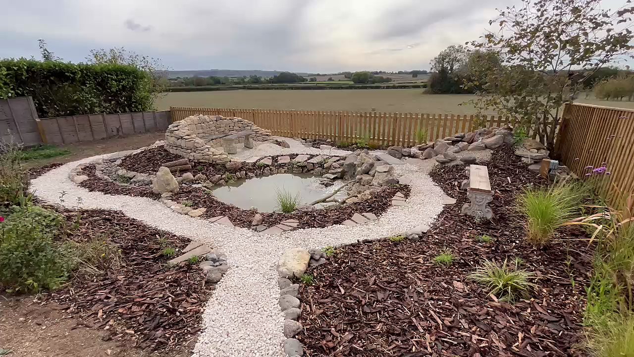 A newly finished wildlife garden: a stone-edged pond, pale gravel paths winding through new planting, and a curved drystone wall with a seat, with open fields beyond the picket fence